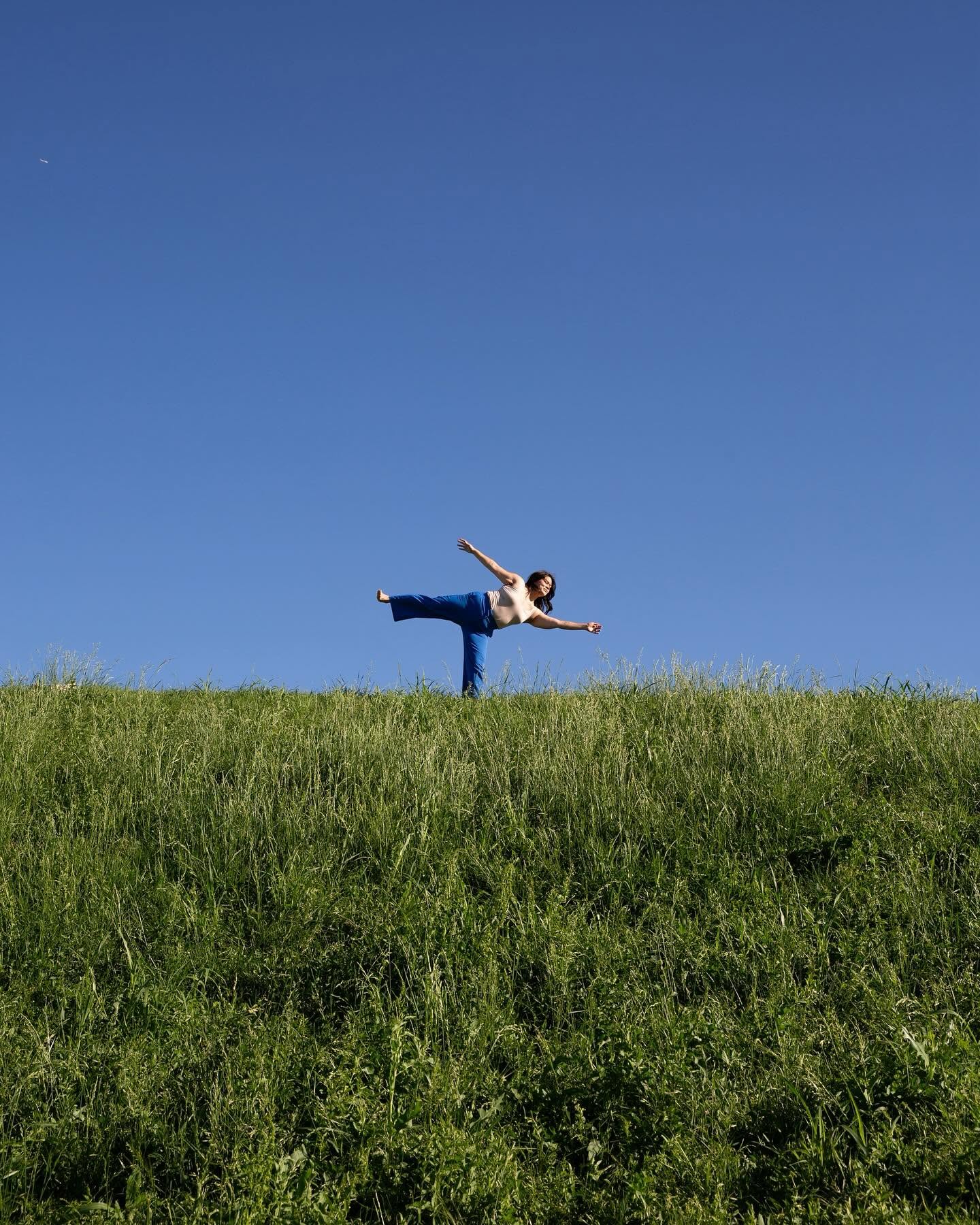 Person balancing on one leg on grassy hill with clear blue sky background.