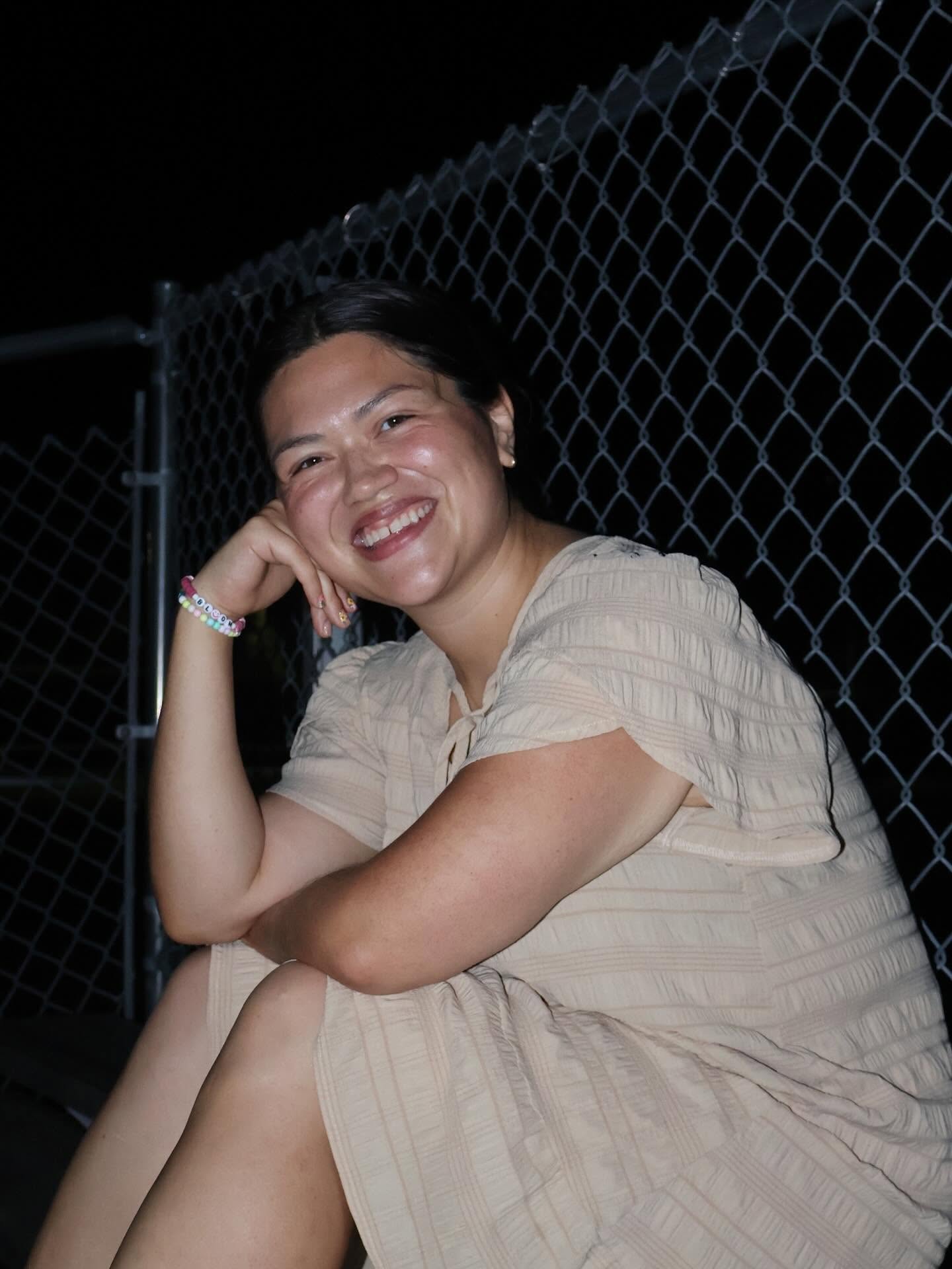 Smiling woman sitting outdoors at night near a chain-link fence, resting her head on her hand, wearing a beige dress.