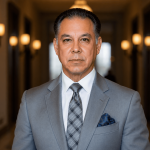 A man in a gray suit and tie standing in a hallway with warm lighting and blurred background.