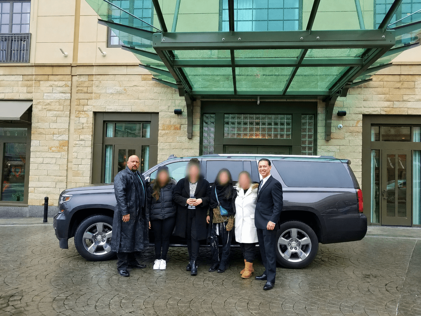 Group of five people standing in front of a black limousine outside a building with glass and stone facade.