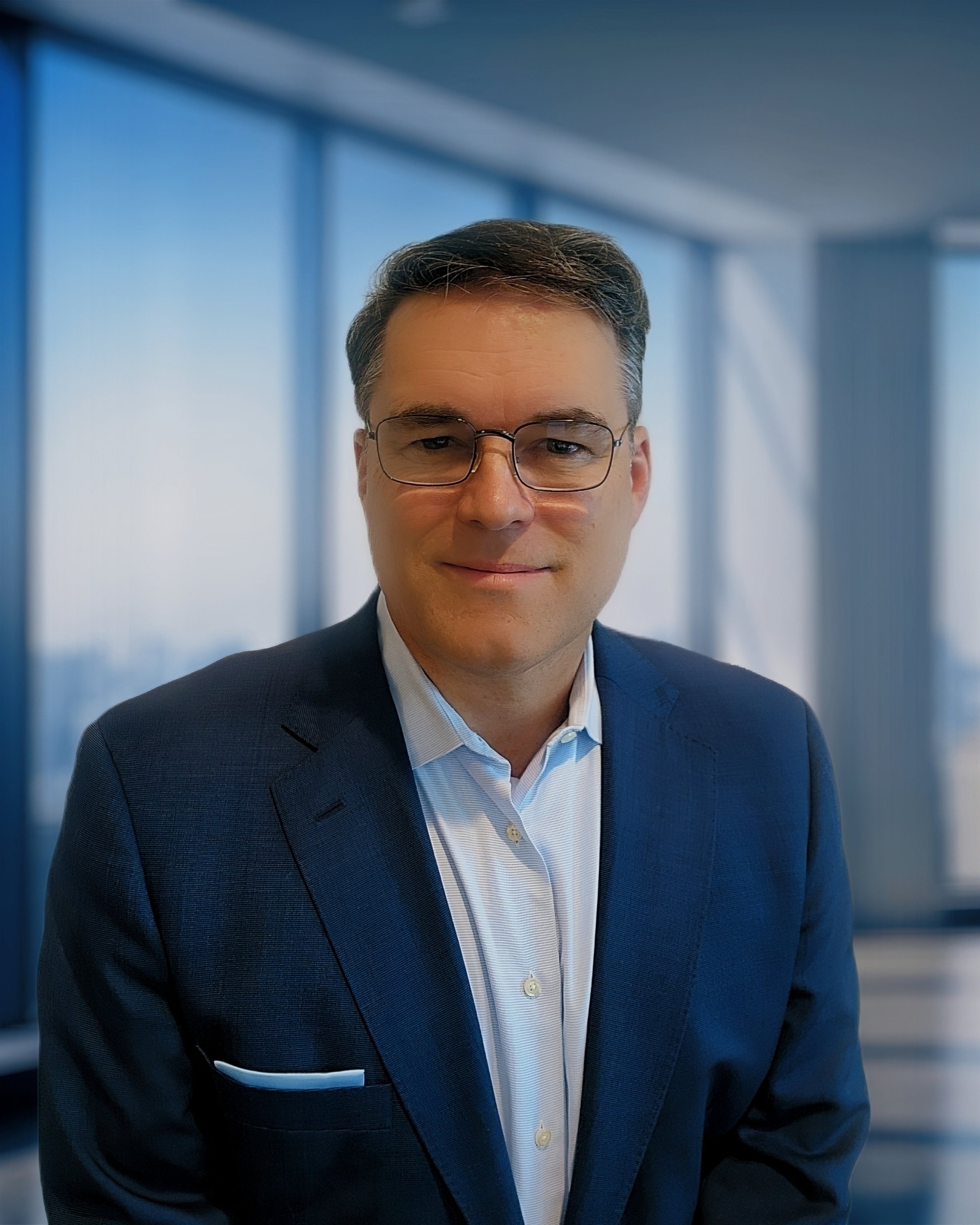 Man in a suit and glasses smiling in a modern office with large windows.