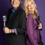 A man and woman stand back-to-back, each holding a vintage microphone, smiling against a purple background.