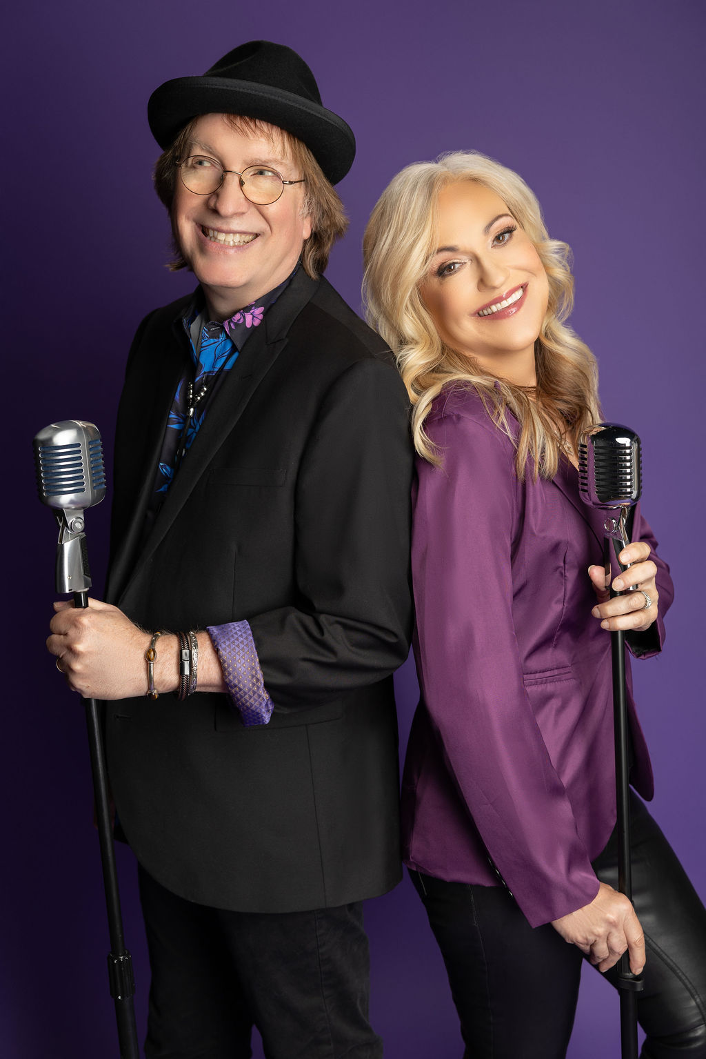 A man and woman stand back-to-back, each holding a vintage microphone, smiling against a purple background.