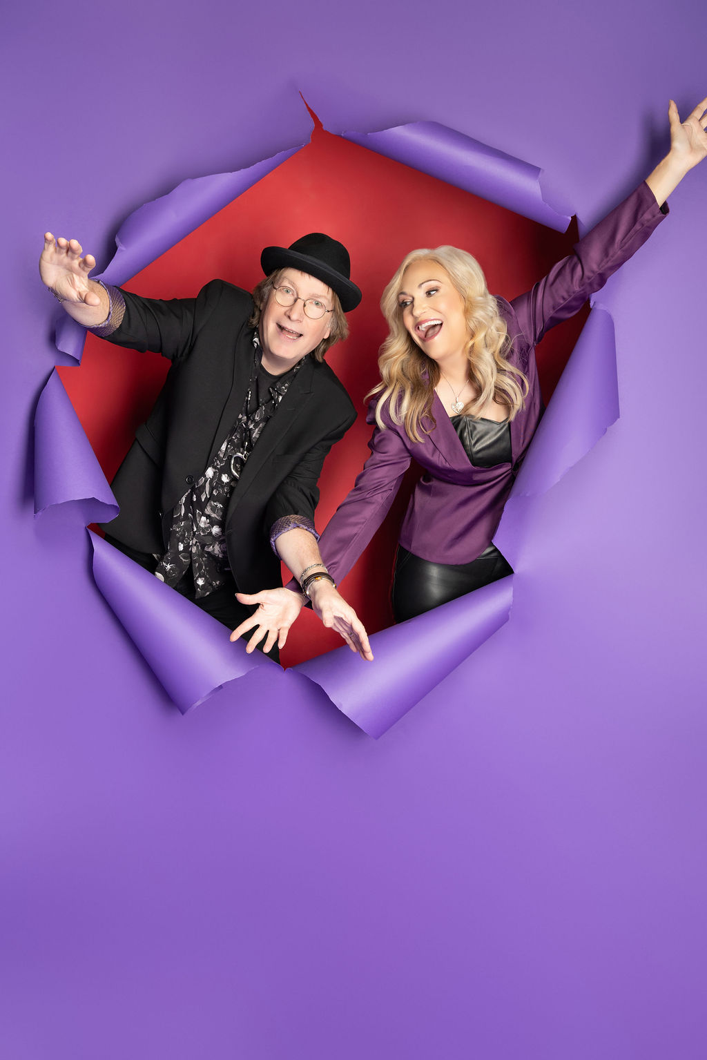 Two people, a man and a woman, smile and wave through a torn purple paper backdrop with a red interior.
