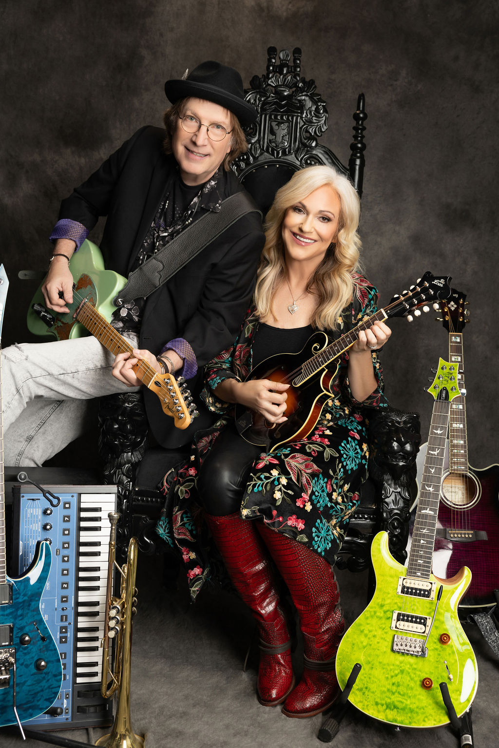 Two smiling women holding guitars, sitting on a decorative chair, surrounded by musical instruments, with a dark background.