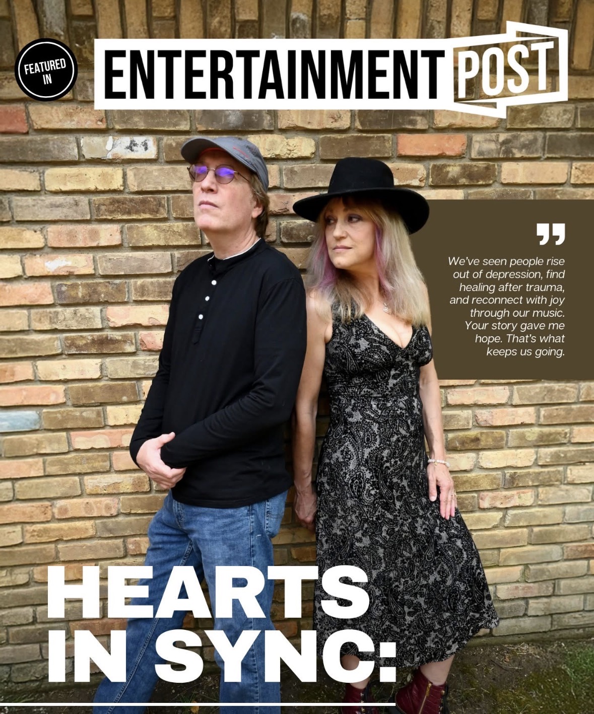 Two people stand against a brick wall, one wearing a hat and glasses, the other in a dress and hat, looking away.