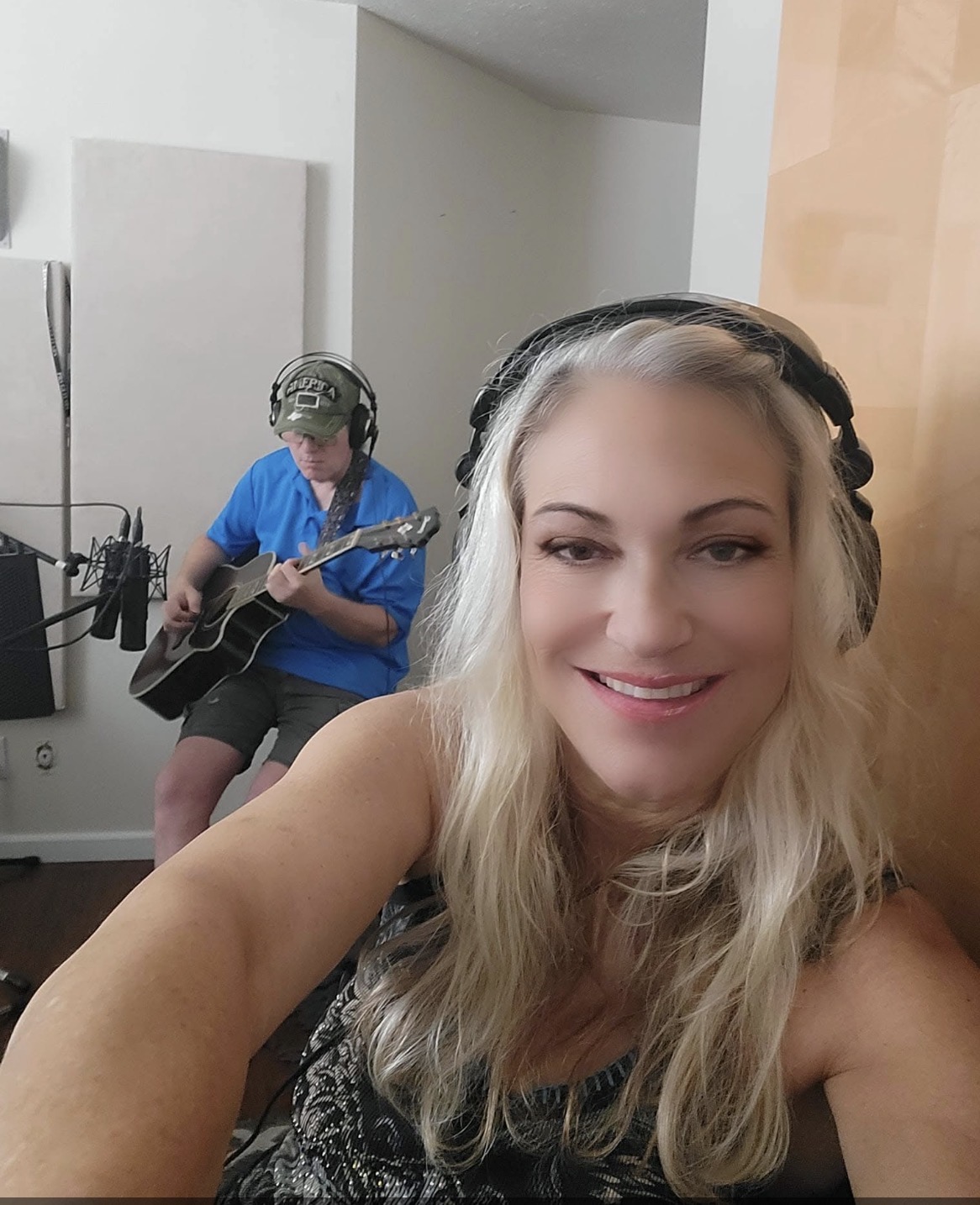 A woman with long blonde hair smiling at the camera, wearing headphones, in a recording studio. A person in blue shirt plays guitar in the background.