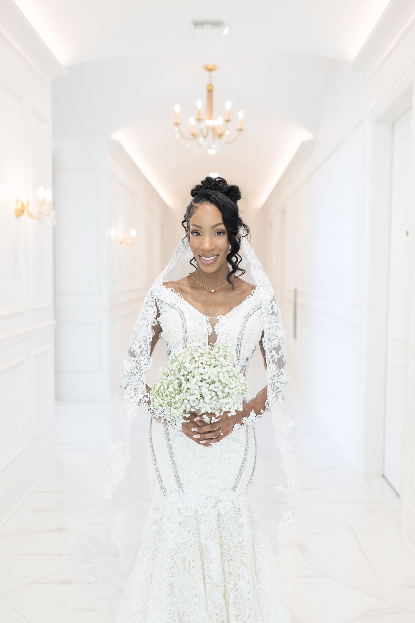 Bride in white lace dress holding bouquet in bright, elegant hallway with chandelier and wall sconces.