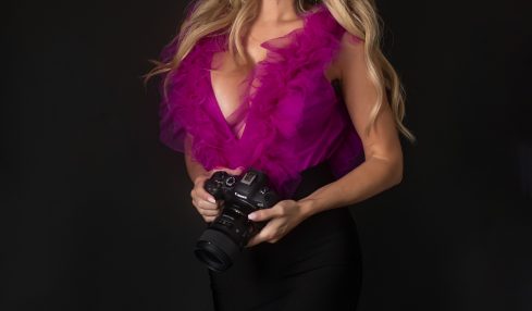 Woman with long blonde hair in a pink feathered top and black skirt holding a camera, smiling, against a black background.