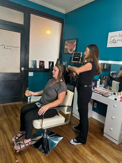Two women in a hair salon, one sitting and the other standing, with salon equipment and a mirror in the background.