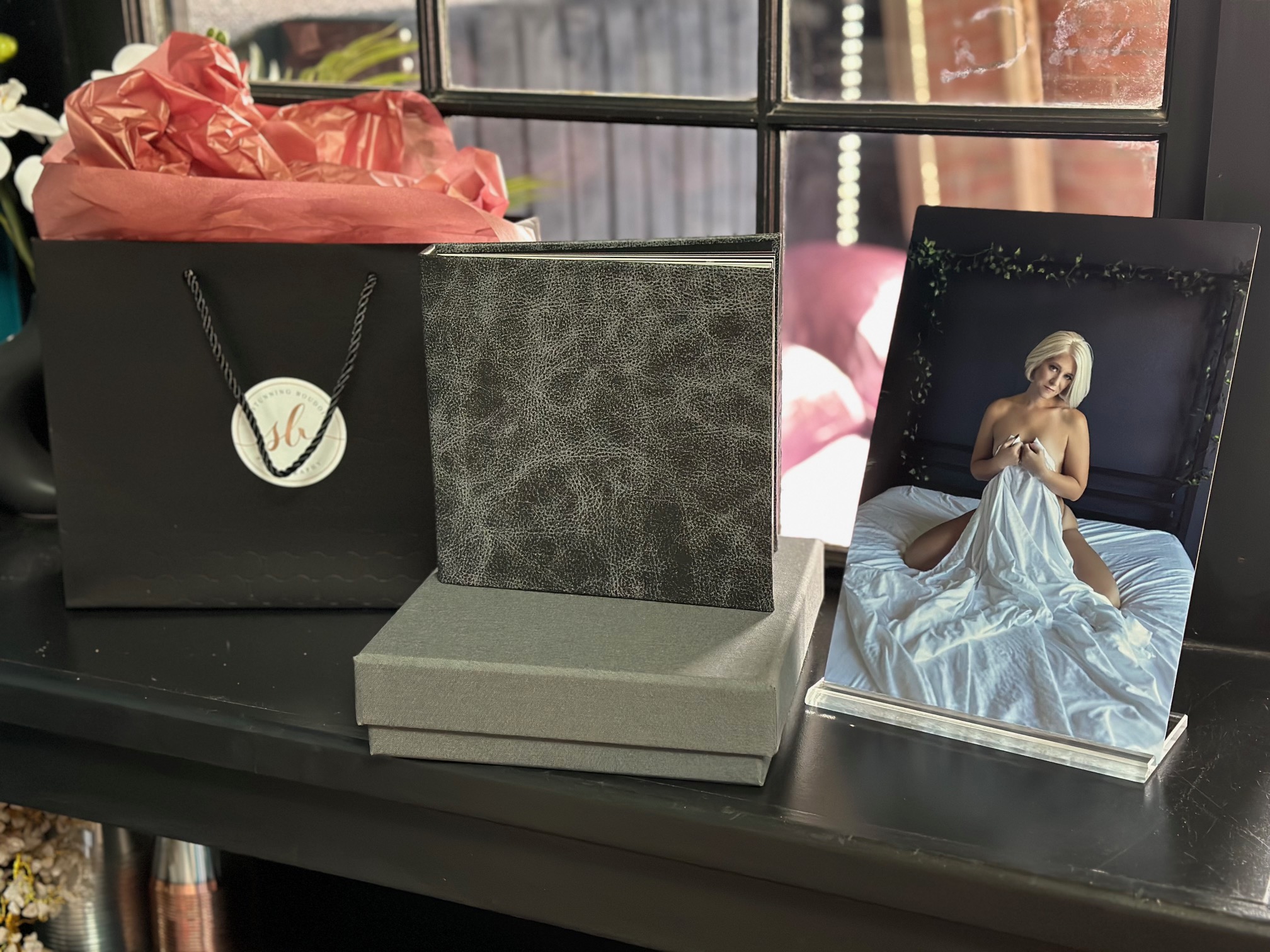 A framed photo of a woman in a white dress sitting on a bed, with a gift bag and box on a black surface near a window.