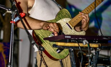 Person playing an acoustic guitar on stage with microphone, wearing a cap and sleeveless shirt.