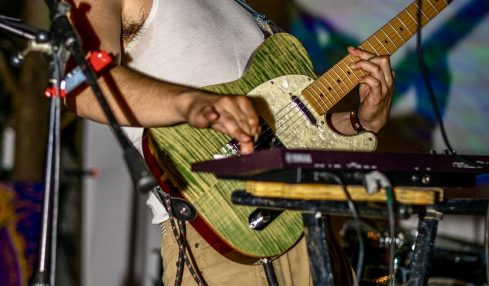 Person playing an acoustic guitar on stage with microphone, wearing a cap and sleeveless shirt.