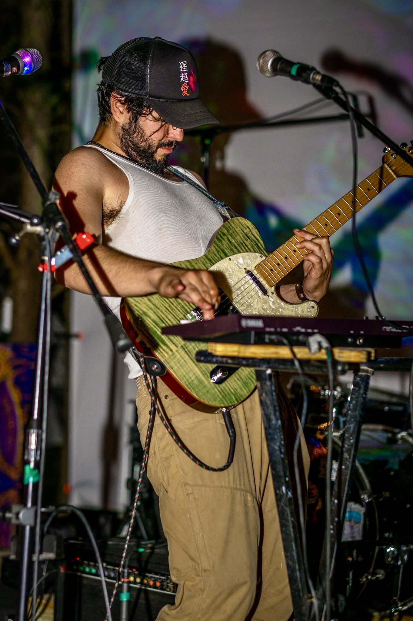 Person playing an acoustic guitar on stage with microphone, wearing a cap and sleeveless shirt.