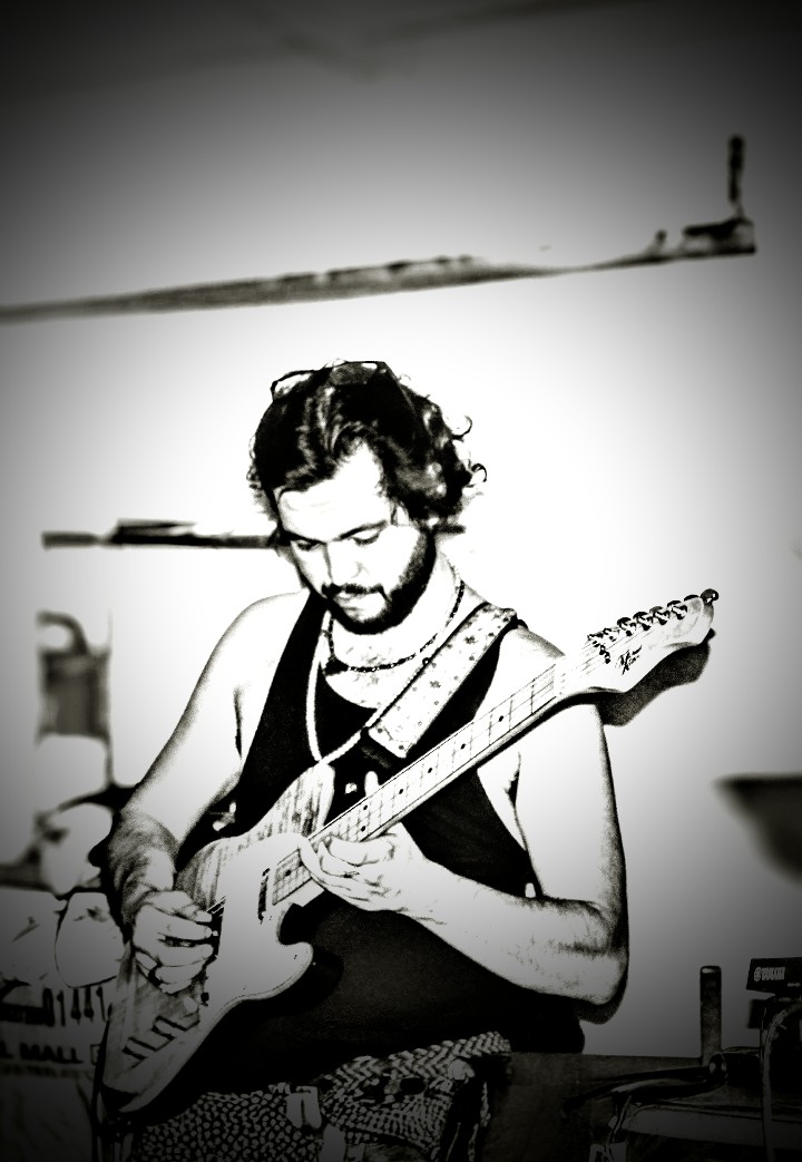 Person with curly hair and beard playing an electric guitar indoors, wearing a sleeveless top and necklaces.
