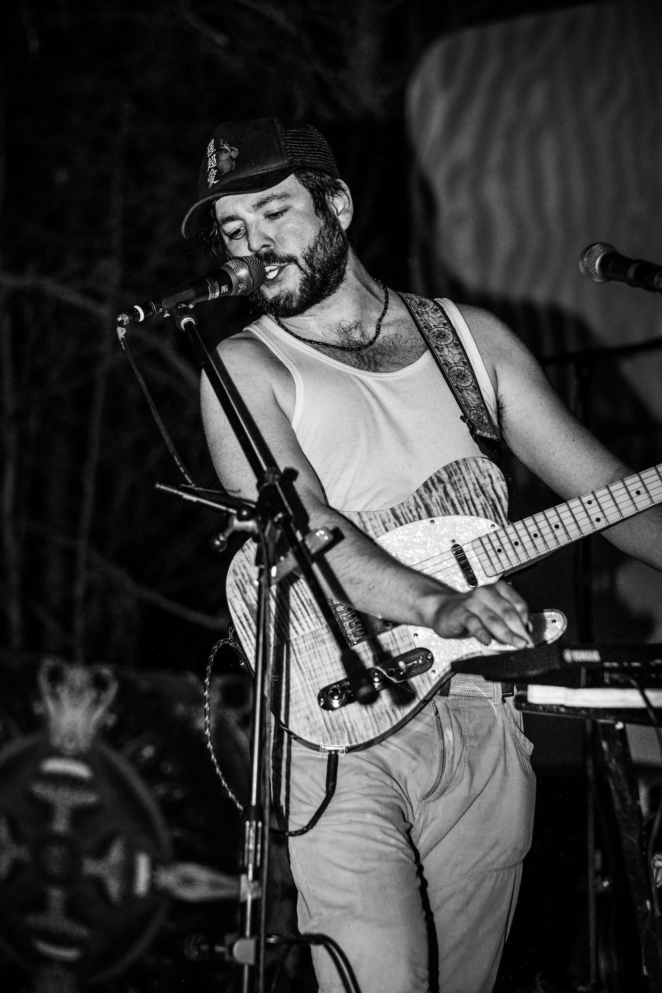 Man with a beard and hat playing guitar and singing into a microphone on stage, smiling, in black and white.