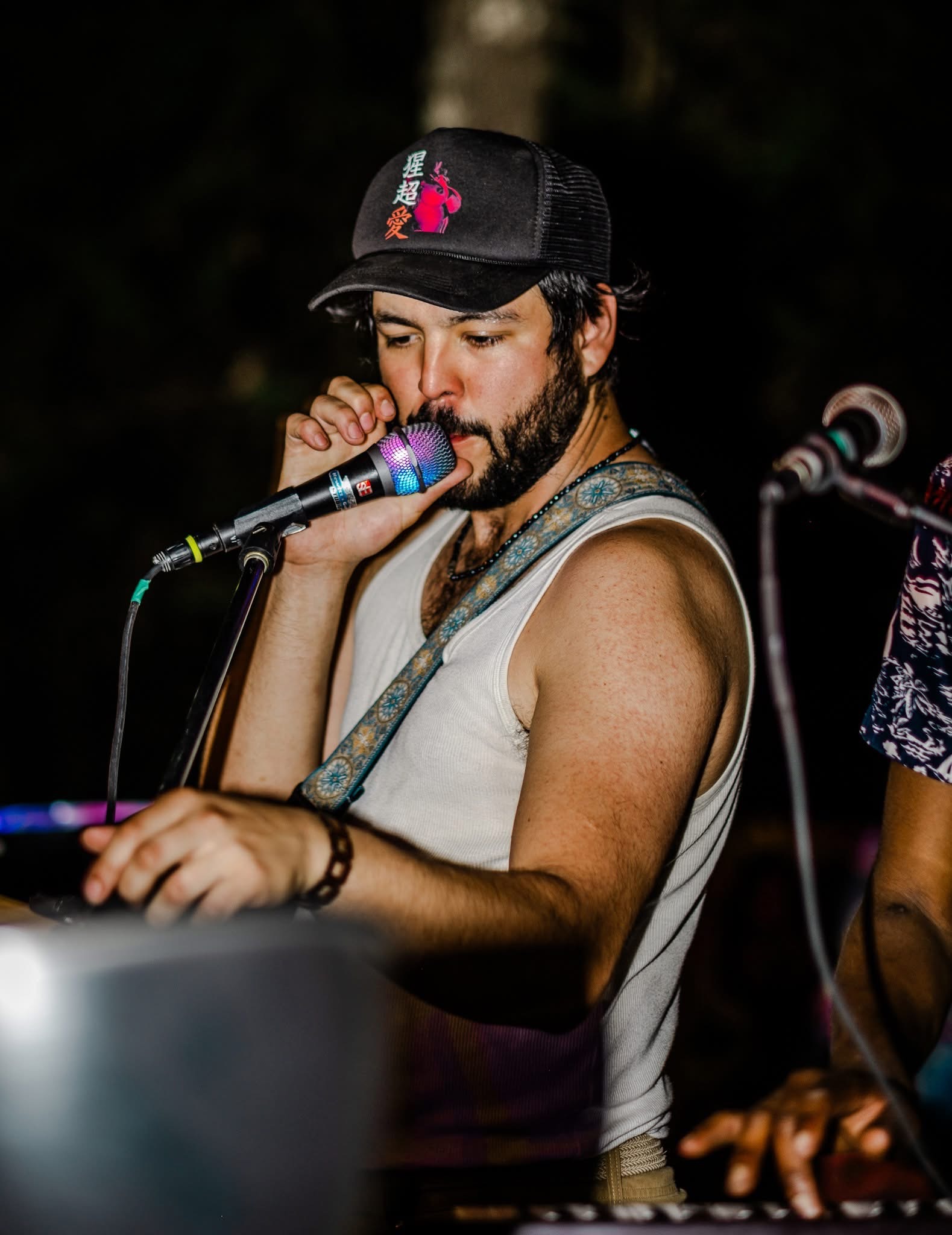 Man with a beard wearing a black cap and sleeveless shirt, singing into a microphone, with a dark background.