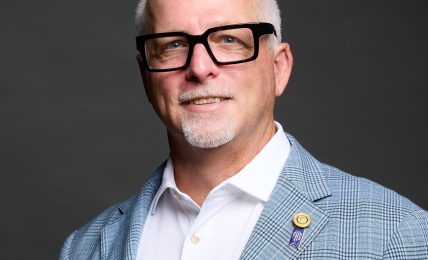 Portrait of a middle-aged man with glasses, wearing a light blue checkered blazer, white shirt, and a purple pocket square, smiling.