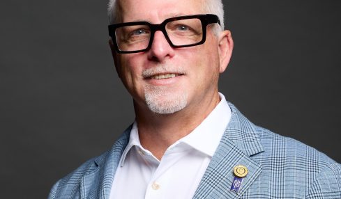 Portrait of a middle-aged man with glasses, wearing a light blue checkered blazer, white shirt, and a purple pocket square, smiling.