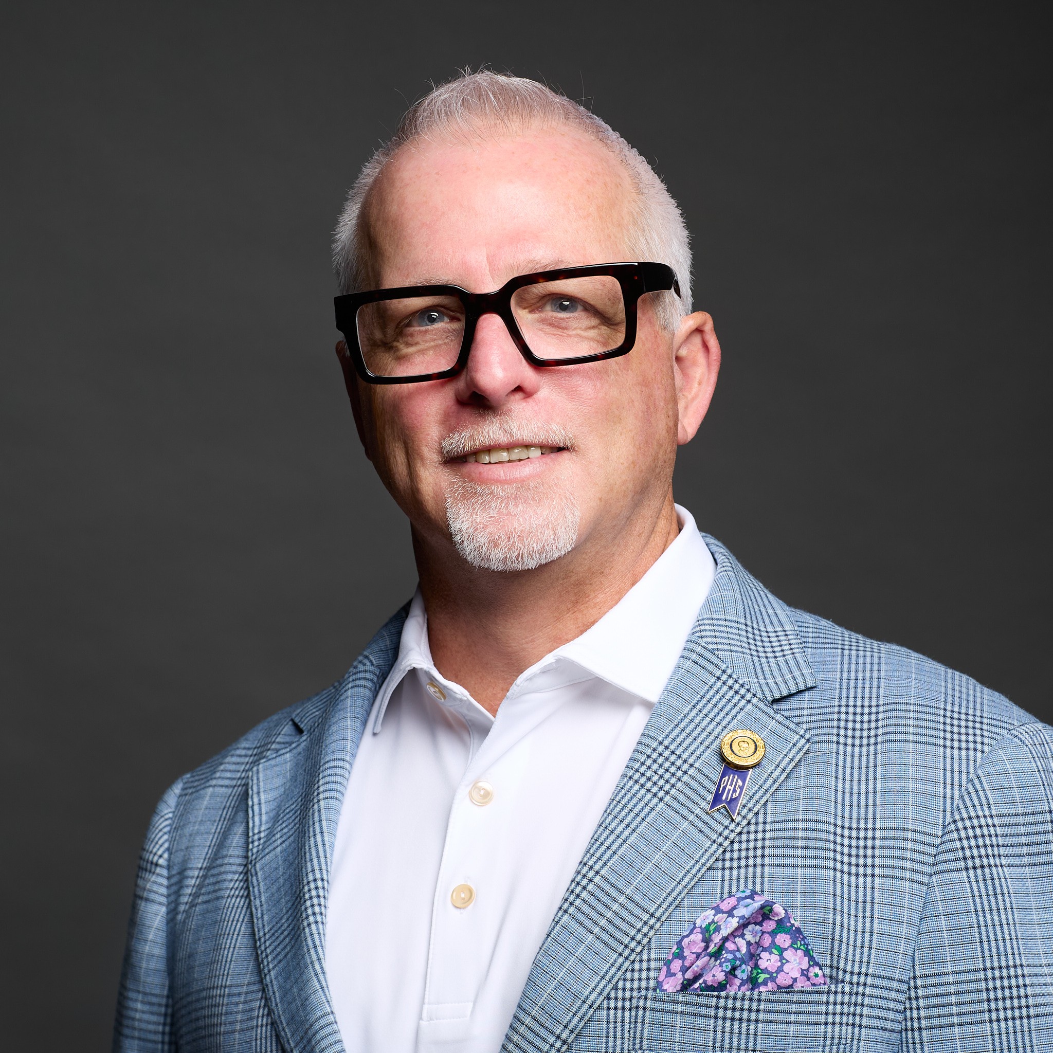 Portrait of a middle-aged man with glasses, wearing a light blue checkered blazer, white shirt, and a purple pocket square, smiling.