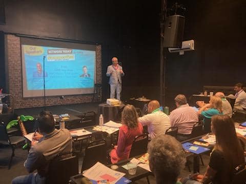 Presenter standing on stage in front of a screen with a slide, audience seated and listening in a conference room.