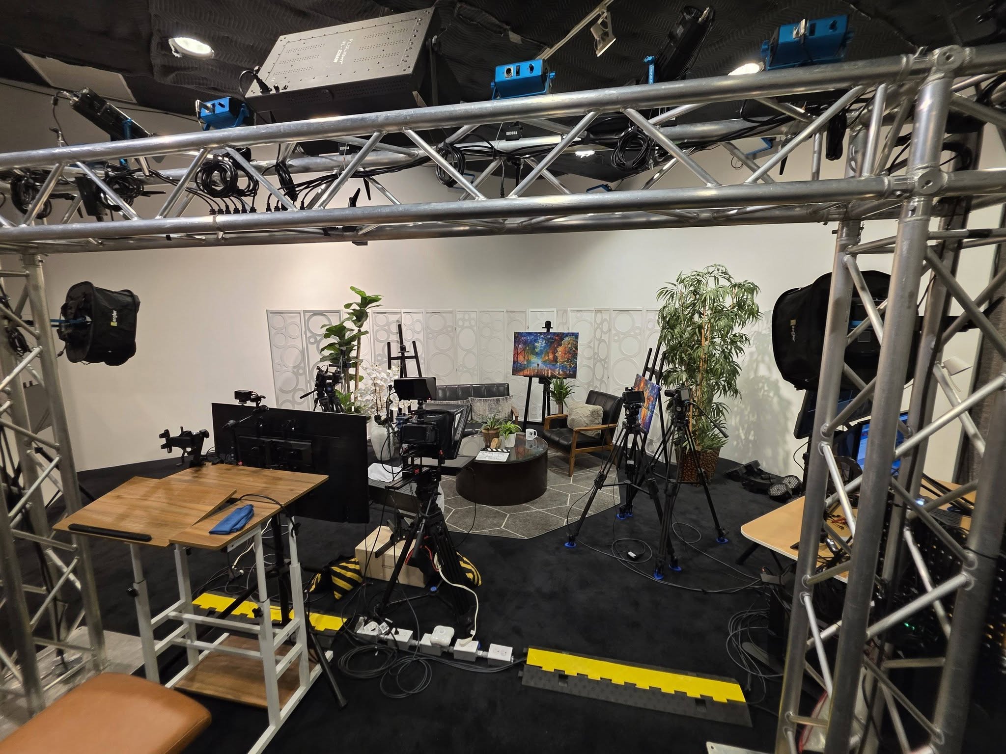 Studio setup with cameras, lighting, and chairs, viewed through metal truss structure, with a white background and plants.