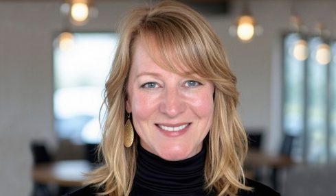 Woman with blonde hair smiling, wearing a black turtleneck, in a modern indoor setting with hanging lights and windows.