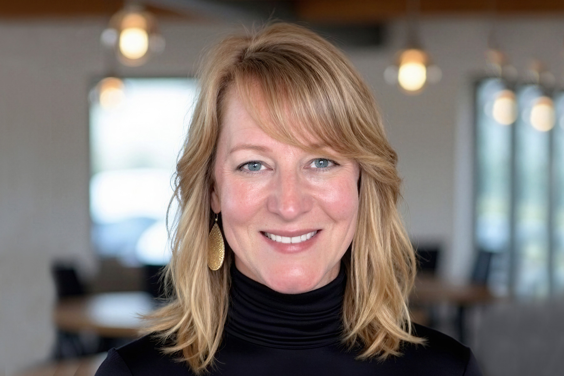 Woman with blonde hair smiling, wearing a black turtleneck, in a modern indoor setting with hanging lights and windows.