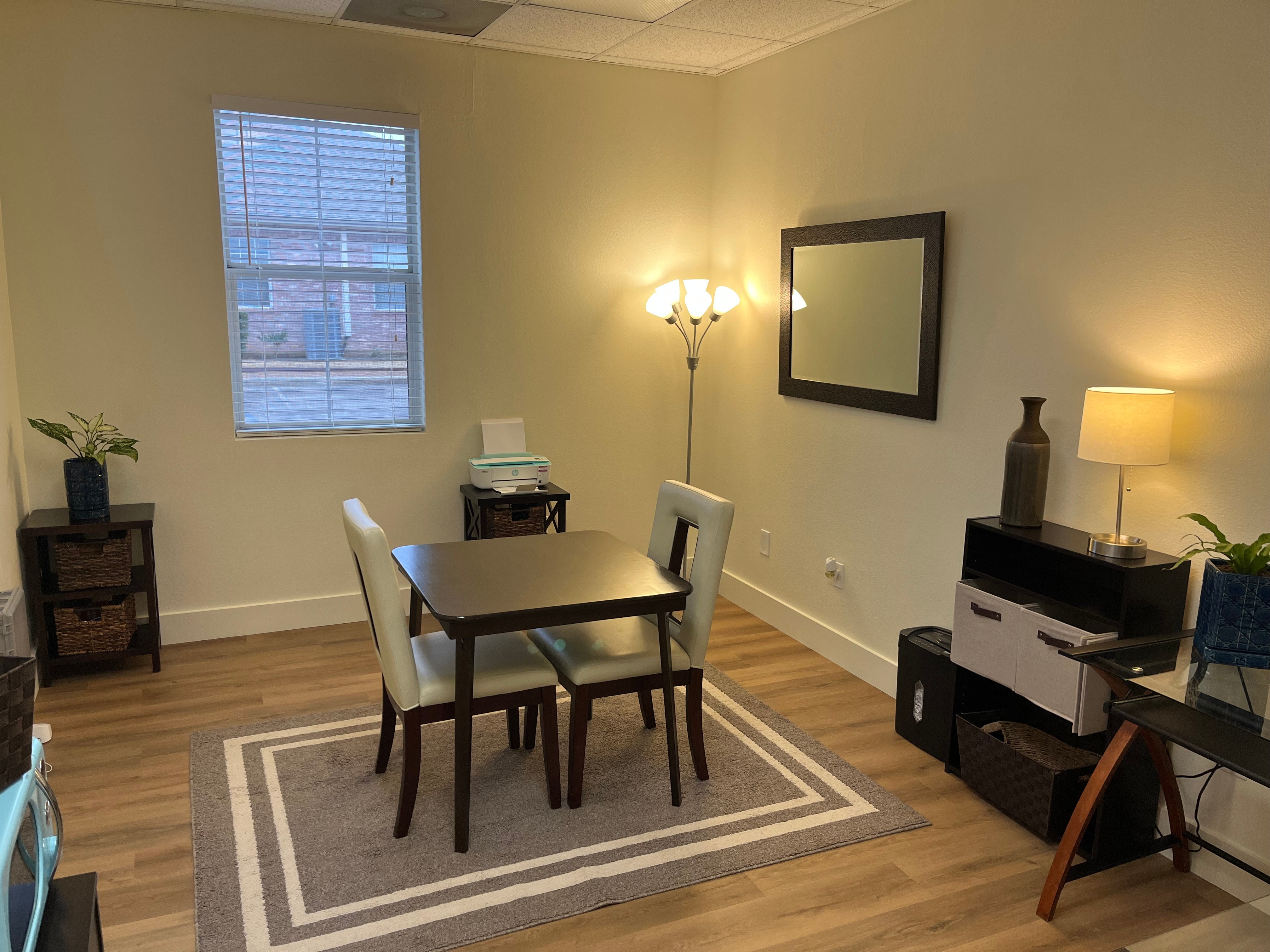 Dining area with table, four chairs, window, mirror, floor lamp, and side table in a room with hardwood floor and beige walls.