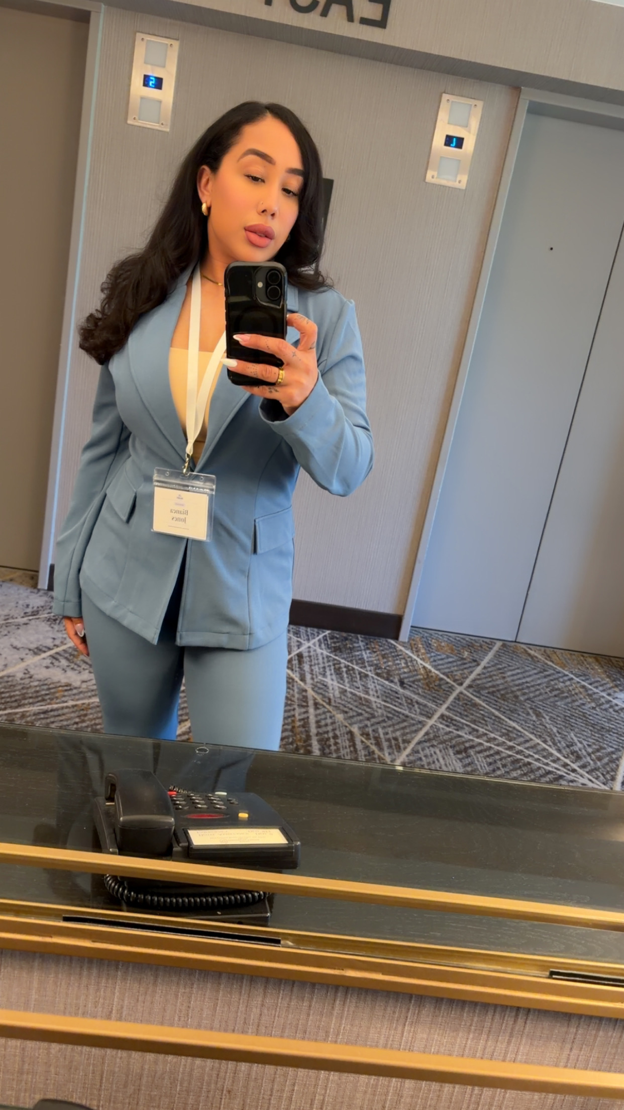Young woman in a light blue suit taking a mirror selfie in an elevator, holding a phone, with a lanyard and badge.