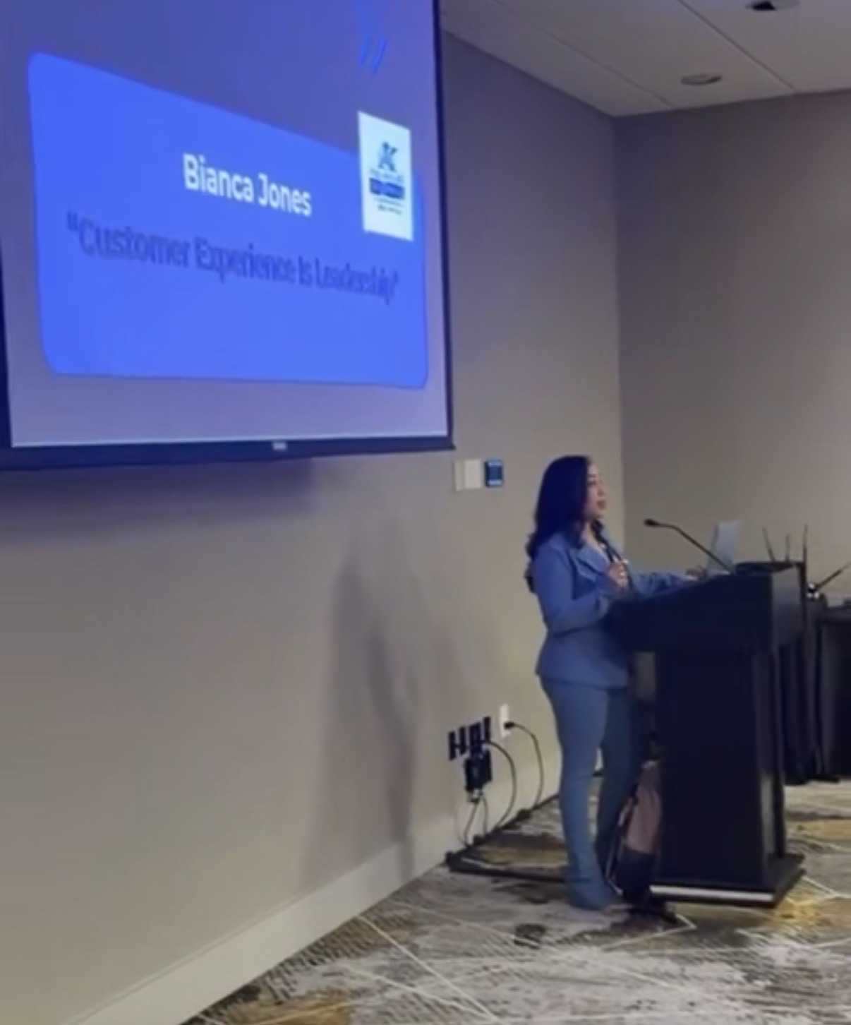 Woman standing at podium giving a presentation in a conference room.