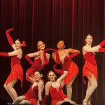 Group of seven dancers in red costumes performing on stage with a dark curtain background.
