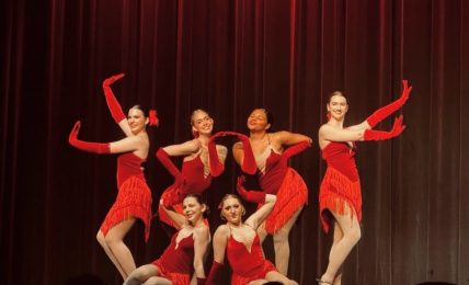 Group of seven dancers in red costumes performing on stage with a dark curtain background.