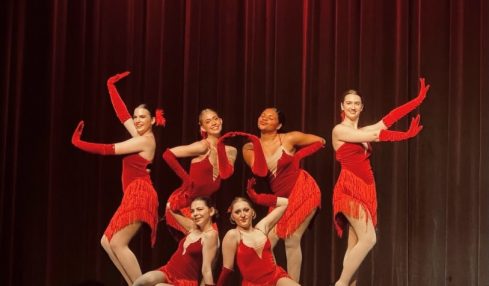 Group of seven dancers in red costumes performing on stage with a dark curtain background.