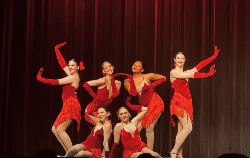 Group of seven dancers in red costumes performing on stage with a dark curtain background.