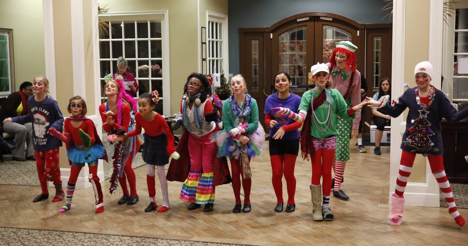 Group of children dancing in a decorated room with large windows and wooden furniture, wearing colorful holiday costumes.