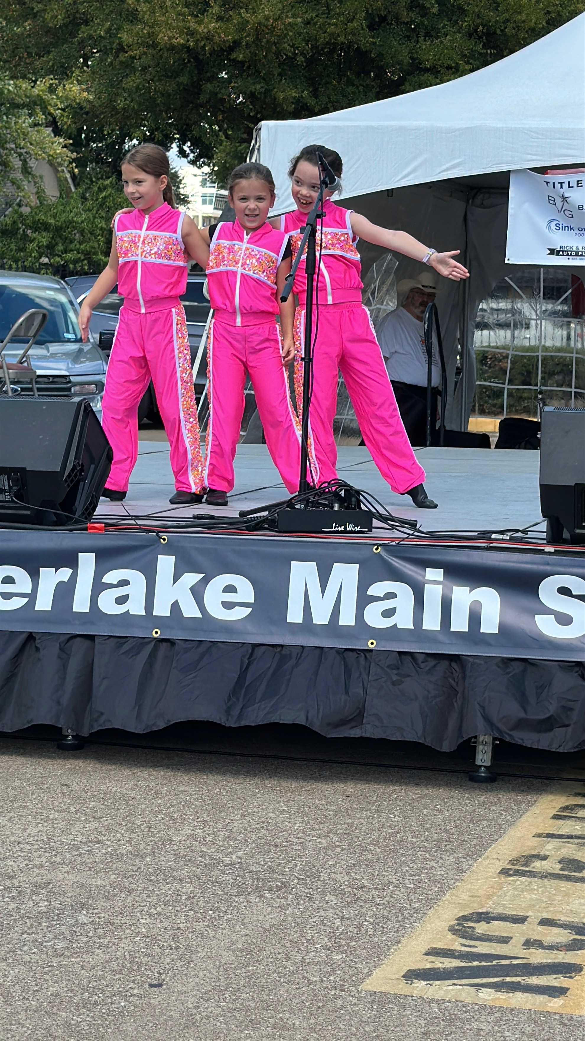 Three young girls in pink outfits perform on an outdoor stage with a tent and trees in the background.