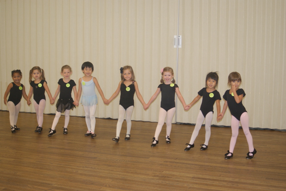 Group of young children holding hands, standing in a line on a wooden floor, in front of a beige curtain.