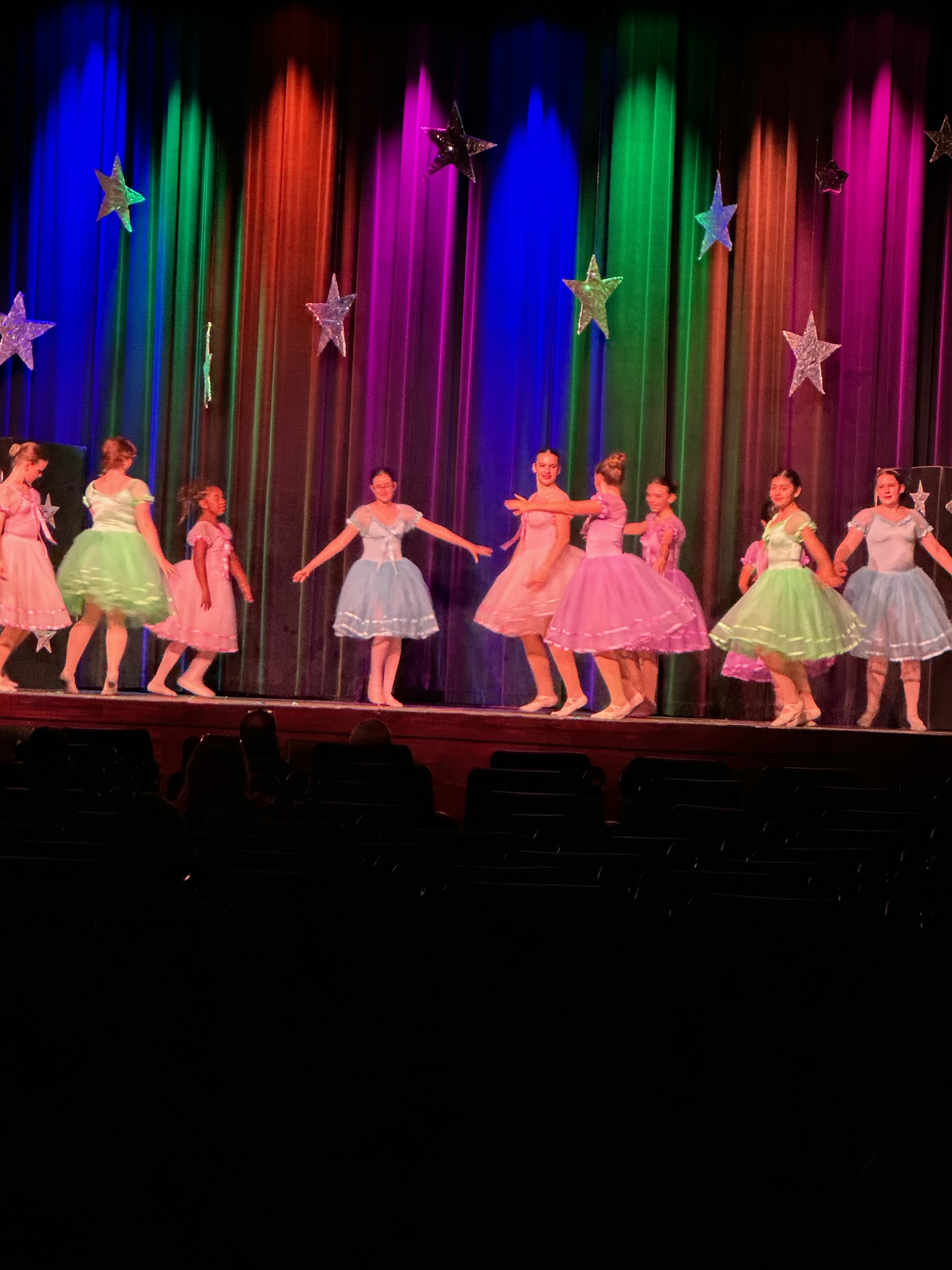 Children in colorful dresses perform on stage with a vibrant, star-decorated curtain backdrop.