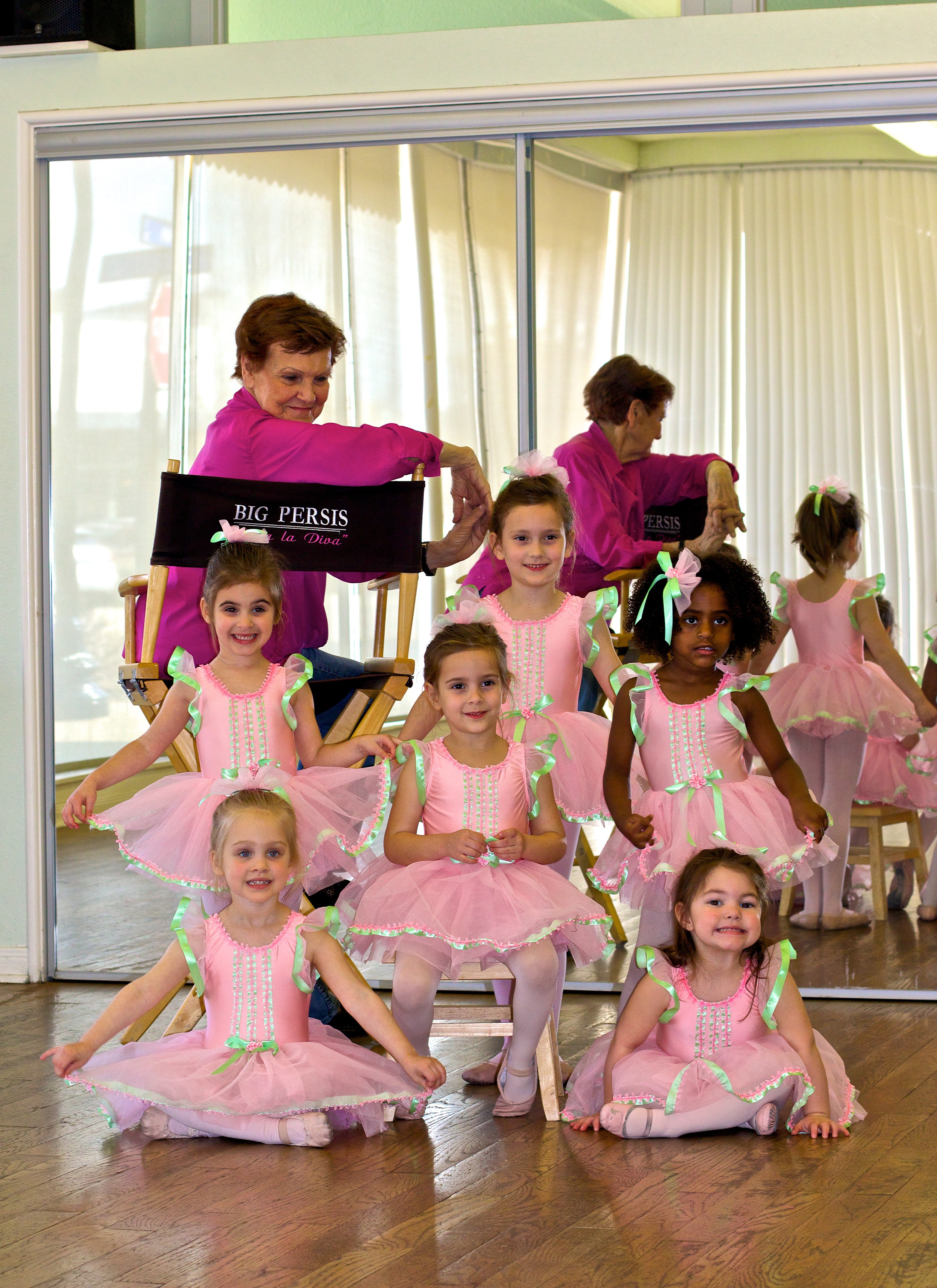 Group of young girls in pink tutus and ballet costumes posing in front of a mirror, with an instructor in the background.
