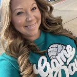 Woman with long wavy hair smiling outdoors, wearing a teal shirt with white text and a graphic, sunny day.