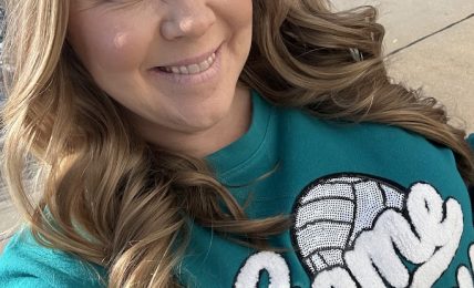 Woman with long wavy hair smiling outdoors, wearing a teal shirt with white text and a graphic, sunny day.