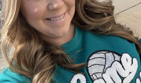 Woman with long wavy hair smiling outdoors, wearing a teal shirt with white text and a graphic, sunny day.