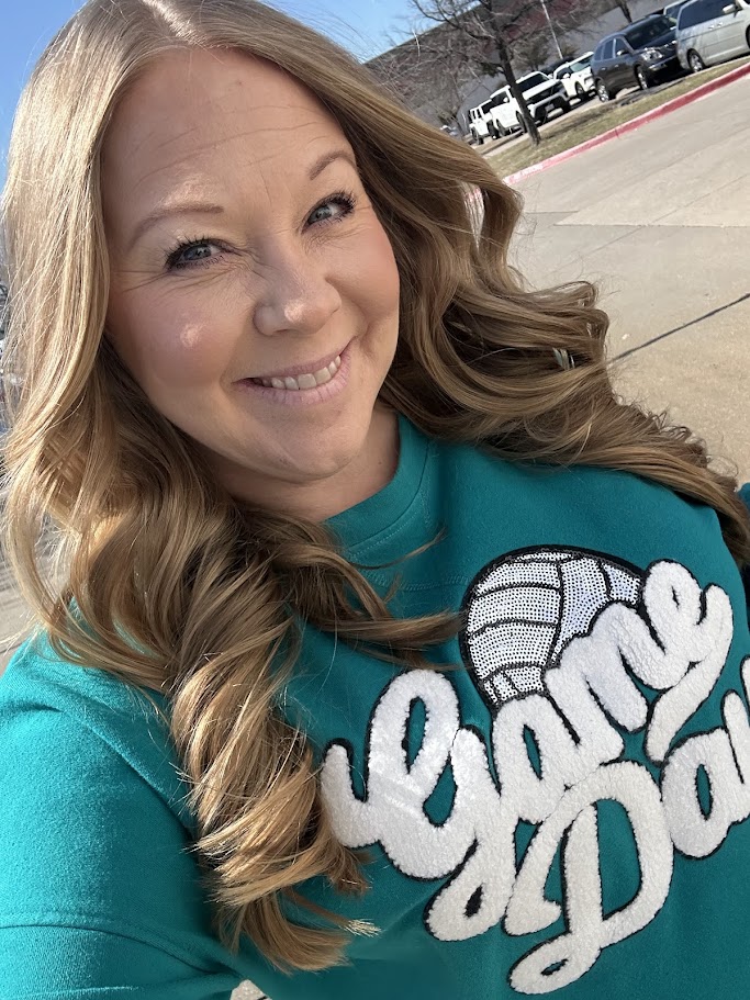 Woman with long wavy hair smiling outdoors, wearing a teal shirt with white text and a graphic, sunny day.