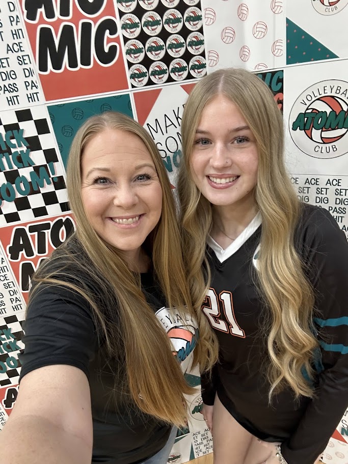 Two smiling women taking a selfie in front of a colorful sports-themed background, one with long straight hair and the other with long wavy hair.