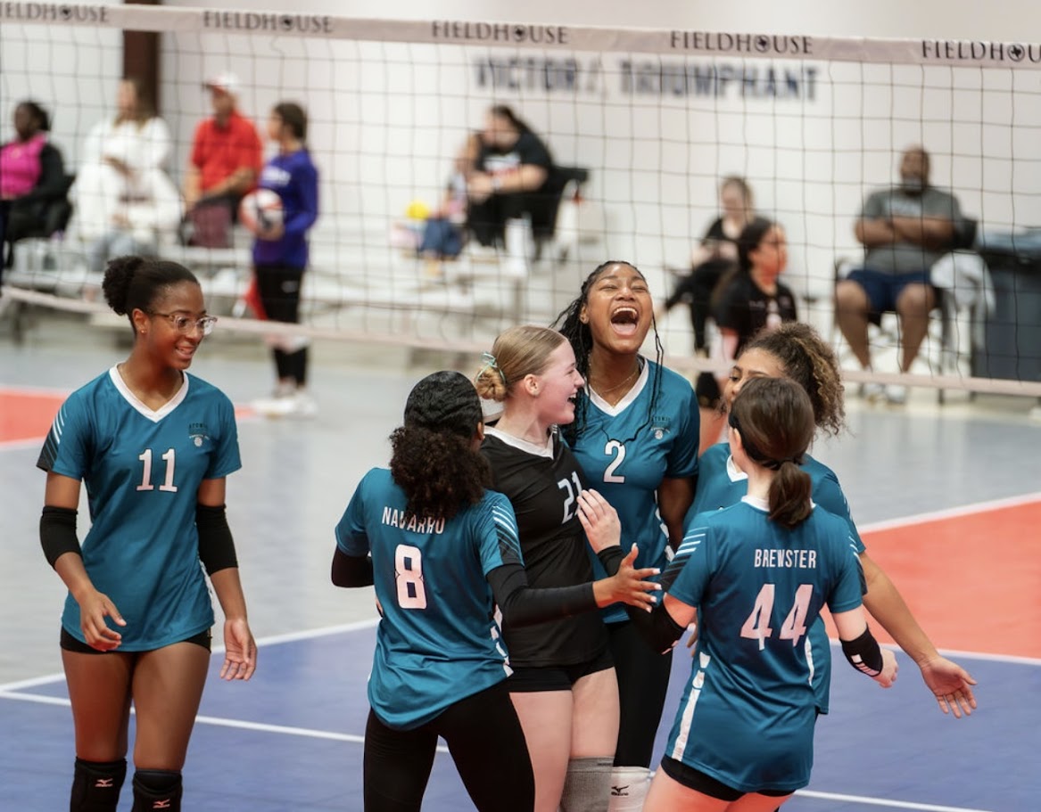 Group of female volleyball players celebrating on court, wearing teal uniforms, with spectators in the background.