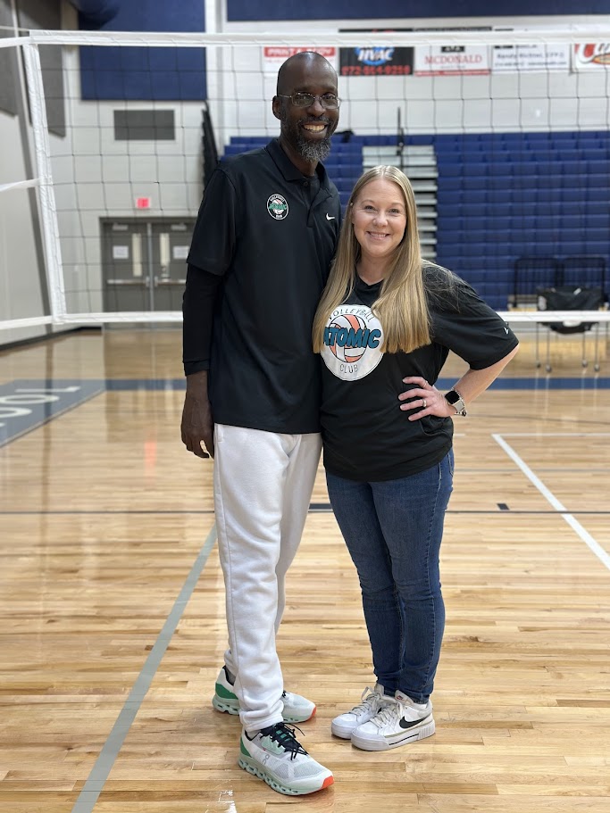 Two people standing on a volleyball court, smiling, with a net and blue bleachers in the background.