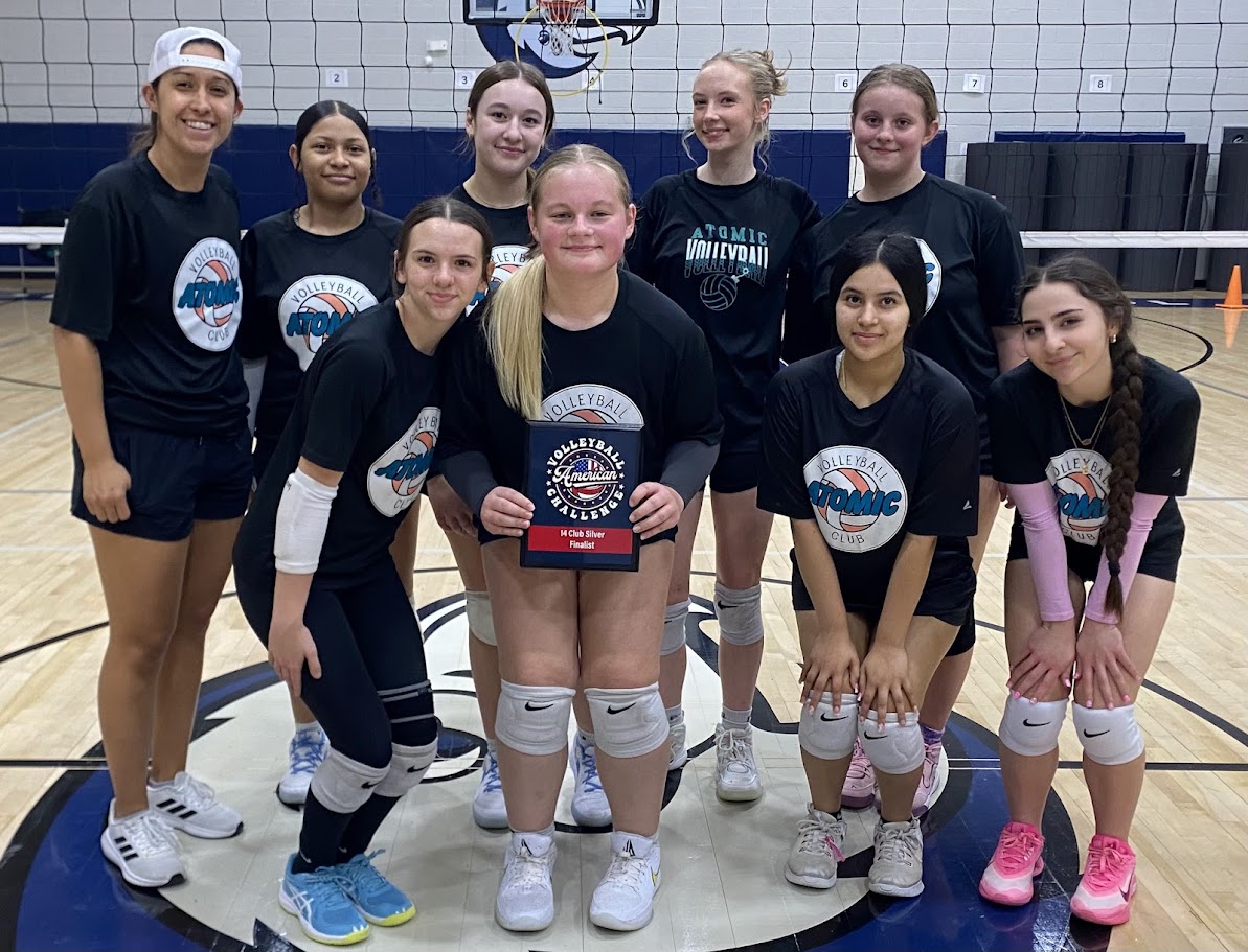 Group of nine young female volleyball players in team uniforms on indoor court, holding a trophy, smiling.