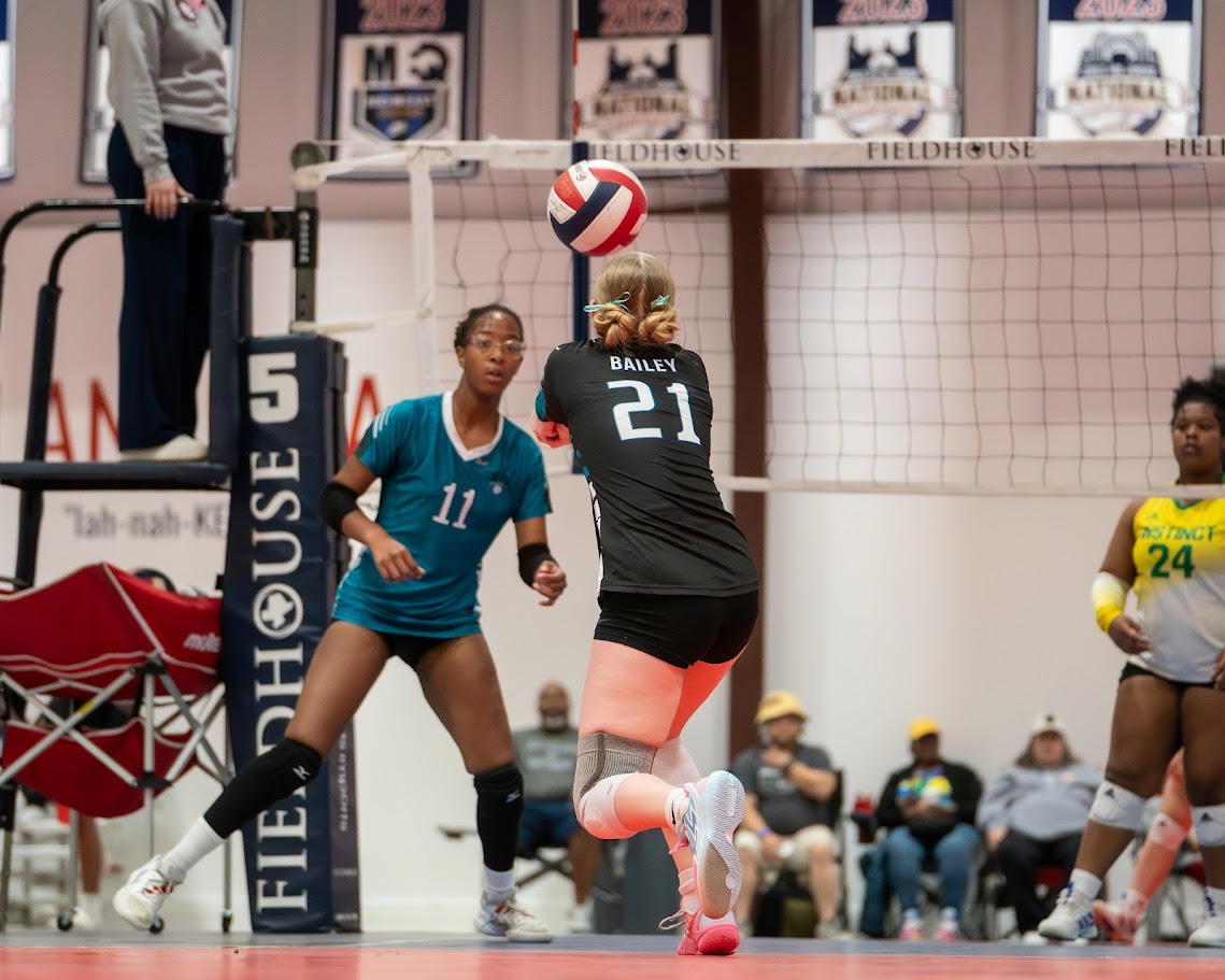 Two female volleyball players near the net, one jumping to hit the ball, others watching, indoor court setting.