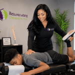 Chiropractor smiling while adjusting a patient's leg during a consultation in a clinic room.